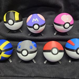 May include: Seven spherical objects, similar to Poké Balls, in various colours including yellow, blue, red, pink, and grey. Each ball has a unique design and sits on a small, black stand. The designs feature geometric patterns and symbols, offering a diverse aesthetic.