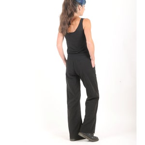 May include: A woman wearing a black tank top and black wide-leg pants. She is standing with her back to the camera and her hands in her pockets.