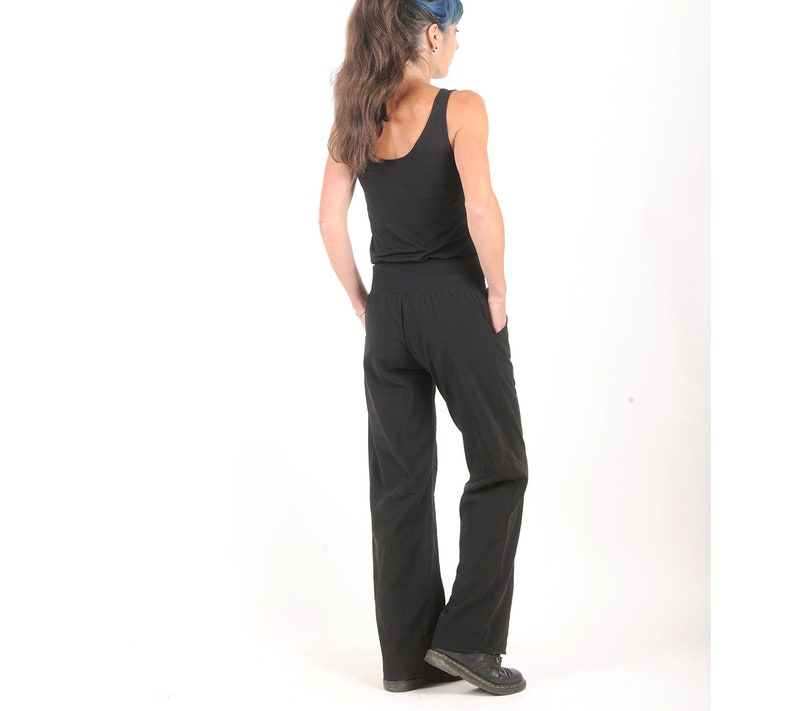May include: A woman wearing a black tank top and black wide-leg pants. She is standing with her back to the camera and her hands in her pockets.
