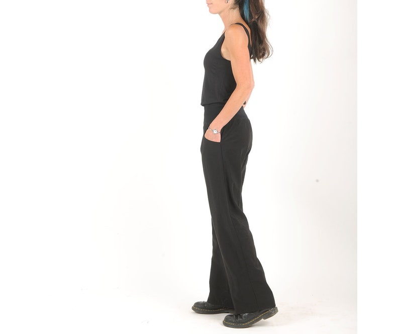 May include: A woman wearing a black tank top and black wide-leg pants. She is standing with her hands in her pockets and looking to the side.