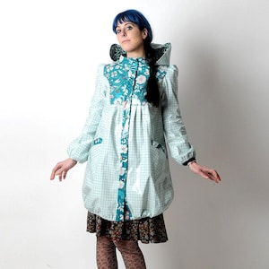 May include: A hooded coat featuring a light blue and white grid pattern with a teal floral yoke and front placket. The coat has long sleeves with elastic cuffs, two side pockets, and a hood lined with a teal and black animal print fabric.