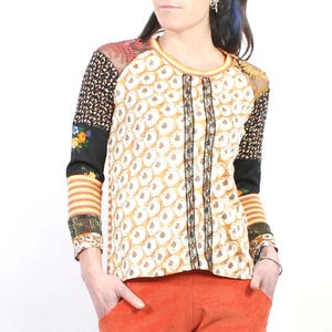Peut inclure: Un haut à manches longues présentant un mélange de motifs, incluant un imprimé floral jaune et blanc sur le devant, des manches noires à motifs floraux et géométriques, et des poignets rayés orange. Le vêtement comporte des détails en dentelle noire.