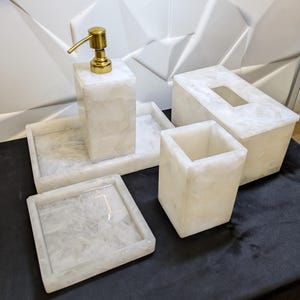 Puede incluir: Conjunto de accesorios de baño de mármol blanco con un dispensador de jabón, una caja de pañuelos, un vaso y dos bandejas. El conjunto tiene un diseño moderno y minimalista.