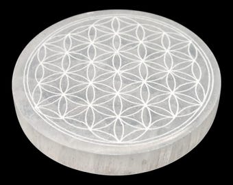 May include: A round, white selenite crystal plate with a carved Flower of Life pattern. The plate is approximately 4 inches in diameter and sits on a black background. The design is a geometric pattern of overlapping circles.