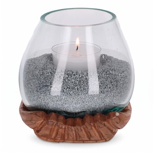 Peut inclure: Un bougeoir en verre transparent posé sur une base en bois brun sculpté. Le bol est rempli de petits cailloux gris et contient une bougie chauffe-plat blanche avec une petite flamme allumée, créant une source de lumière chaleureuse.