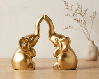 May include: Two gold-colored elephant figurines positioned facing each other with their trunks touching at the tips. The elephants are depicted in a seated posture with smooth, metallic surfaces and detailed ears, eyes, and trunks.