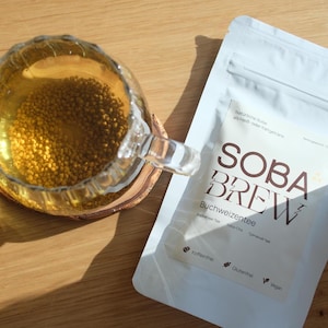 Könnte beinhalten: Ein weißer Beutel mit dem Text "SOBA BREW" in braunen Buchstaben. Der Beutel enthält Buchweizen-Tee, auch bekannt als Soba-Cha. Der Text "Buchweizentee" steht unter dem Markennamen. Der Beutel ist als koffeinfrei, glutenfrei und vegan gekennzeichnet.