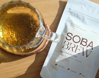 Puede incluir: Una bolsa blanca con el texto "SOBA BREW" en letras marrones. La bolsa contiene té de trigo sarraceno, también conocido como soba-cha. El texto "Buchweizentee" está escrito debajo del nombre de la marca. La bolsa está etiquetada como sin cafeína, sin gluten y vegana.