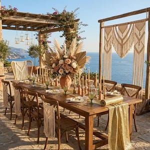 May include: A beautifully set outdoor dining table with a wooden frame, adorned with floral arrangements, candles, and elegant place settings. The table is set for a special occasion, with a macrame backdrop and a view of the ocean.
