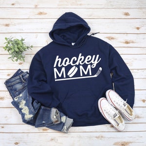 May include: Navy blue hoodie with the words "hockey MOM" in white script and block letters, along with a hockey puck and stick graphic. The hoodie is paired with blue jeans and white sneakers.