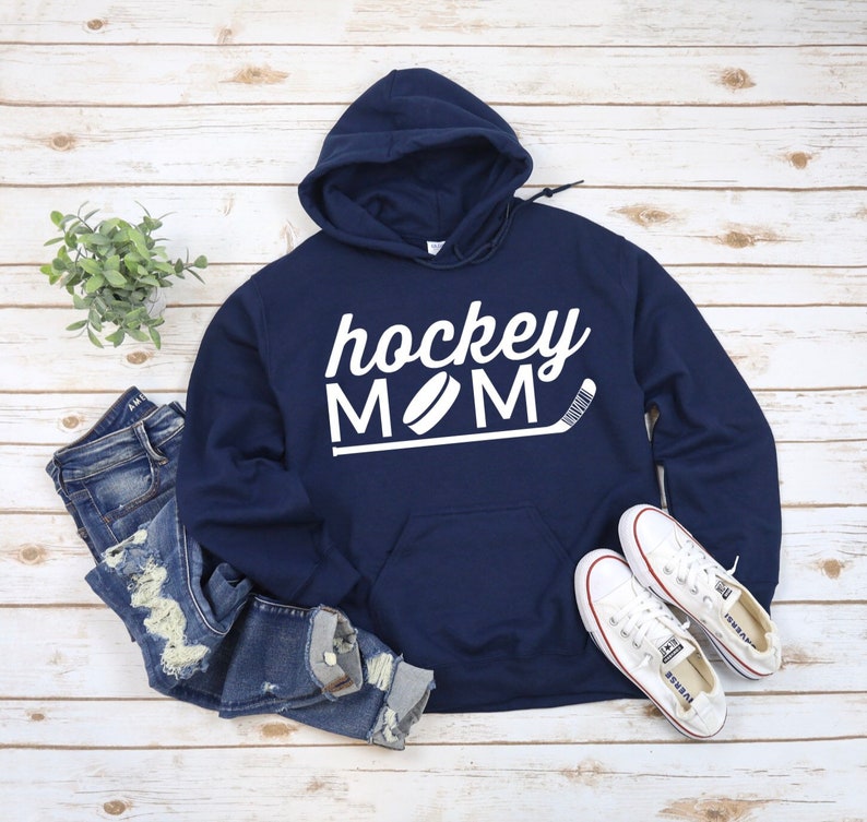 May include: Navy blue hoodie with the words "hockey MOM" in white script and block letters, along with a hockey puck and stick graphic. The hoodie is paired with blue jeans and white sneakers.