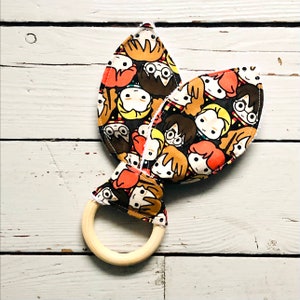 May include: A baby teether with a wooden ring and a fabric design featuring cartoon characters with glasses and brown hair on a black background. The fabric has orange, white, and yellow accents.