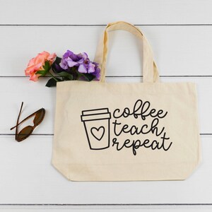 以下が含まれることがあります： 「coffee teach repeat」の黒い文字が書かれたベージュ色のキャンバス地のトートバッグ。左側には、ハートの付いた白黒のコーヒーカップのグラフィックがあります。バッグには2つのハンドルがあり、ピンクと紫の花で満たされています。