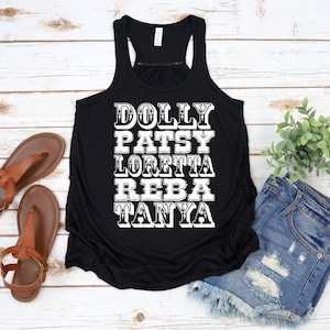 Puede incluir: Camiseta sin mangas negra con texto blanco que enumera los nombres Dolly, Patsy, Loretta, Reba y Tanya. La camiseta se muestra sobre una superficie de madera clara, con sandalias marrones y pantalones cortos de mezclilla en el fondo.