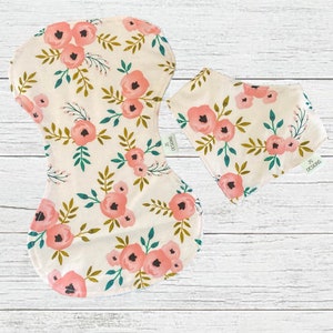 May include: A set of two items: a reusable menstrual pad and a bandana bib. Both are cream-colored and feature a floral pattern with pink roses, green leaves, and brown stems. The bib has a tag that reads "JS DESIGNS".