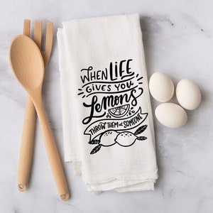 Könnte beinhalten: Weißes Küchentuch mit dem schwarzen Text "When life gives you lemons, throw them at someone". Das Tuch zeigt eine Zitronenscheibe und zwei Zitronen. Holzlöffel, Gabel und drei weiße Eier sind ebenfalls im Bild.