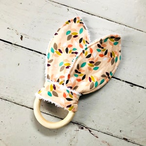 May include: A baby rattle shaped like bunny ears. The ears are made of fabric with a beige background and a colorful leaf pattern. The ears are attached to a natural wood ring. The edges of the ears have a white, fringed trim.