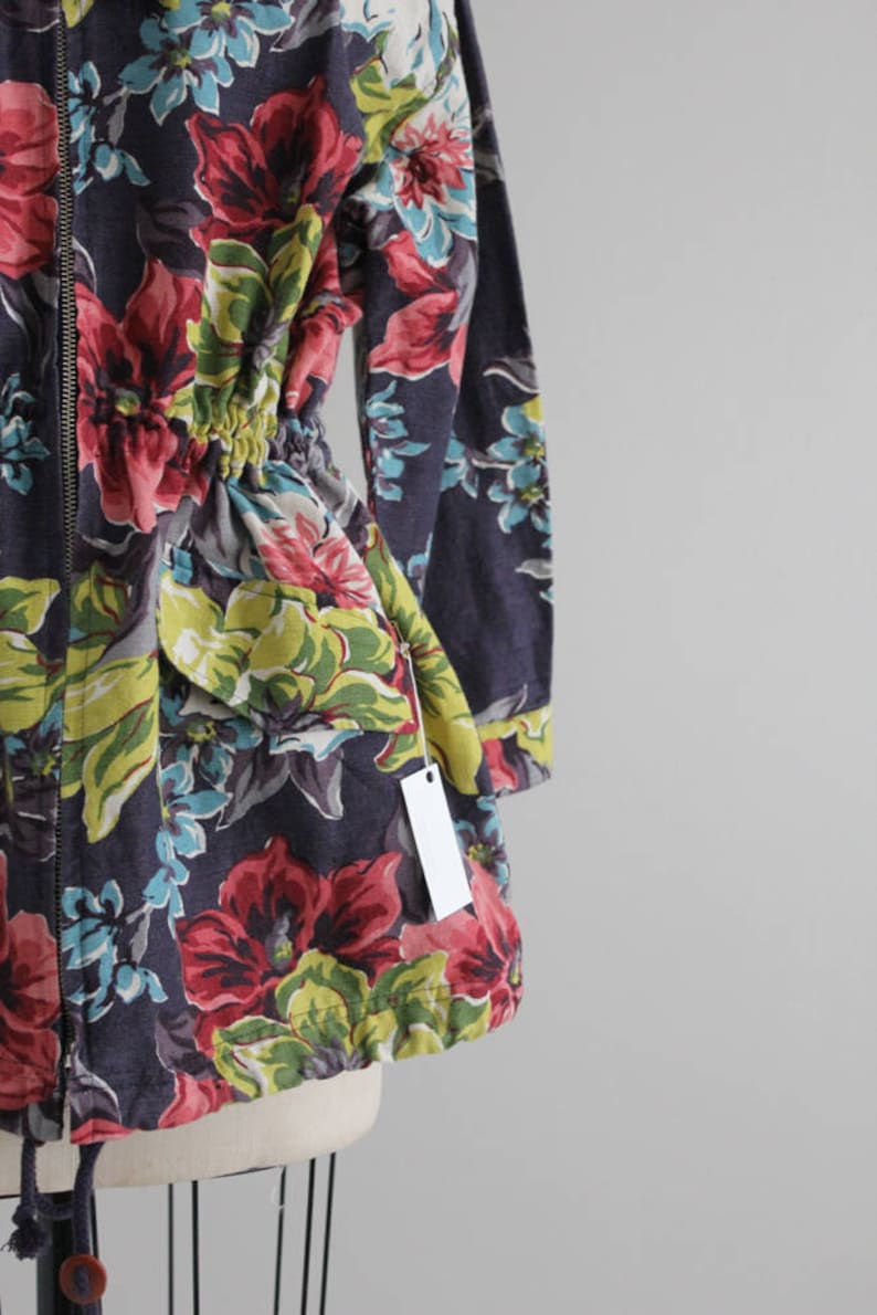 May include: A dark blue floral print jacket with a zipper closure and a drawstring waist. The jacket has a tag attached to the sleeve.