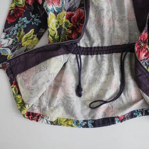 May include: A purple and floral patterned jacket with a drawstring waist and a zipper closure. The jacket is lined with a light gray fabric.
