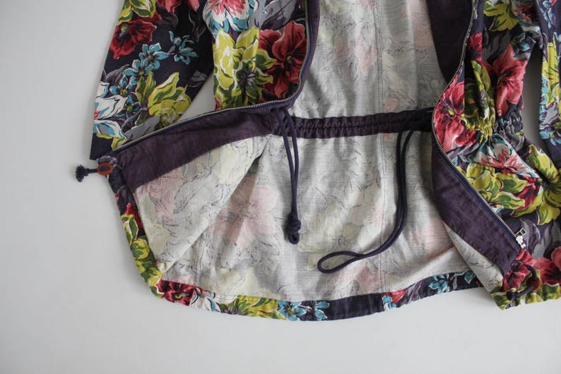 May include: A purple and floral patterned jacket with a drawstring waist and a zipper closure. The jacket is lined with a light gray fabric.
