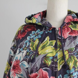 May include: A hooded sweatshirt with a floral print in shades of pink, blue, green, and white on a dark blue background. The zipper is closed.