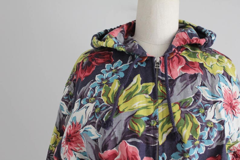 May include: A hooded sweatshirt with a floral print in shades of pink, blue, green, and white on a dark blue background. The zipper is closed.