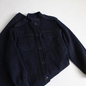 May include: A navy blue jacket with a stand-up collar, long sleeves, and multiple pockets. The jacket has a button closure.