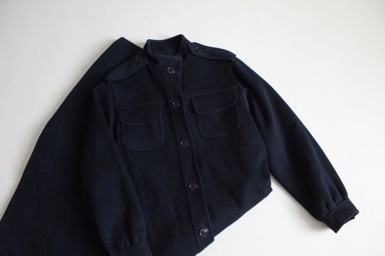 May include: A navy blue jacket with a stand-up collar, long sleeves, and multiple pockets. The jacket has a button closure.