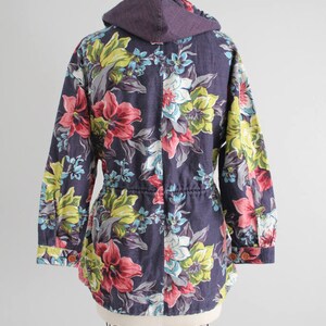 May include: A purple hooded jacket with a floral print featuring pink, yellow, and blue flowers. The jacket has a drawstring waist and long sleeves.