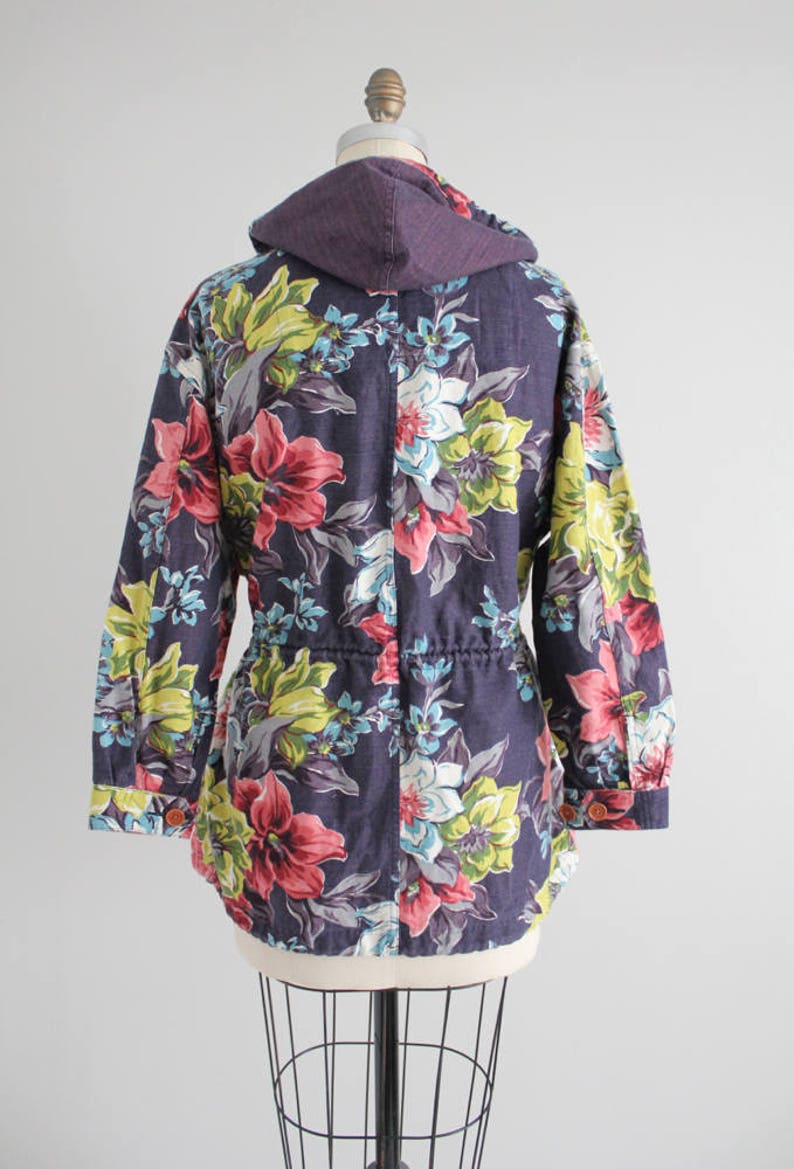 May include: A purple hooded jacket with a floral print featuring pink, yellow, and blue flowers. The jacket has a drawstring waist and long sleeves.
