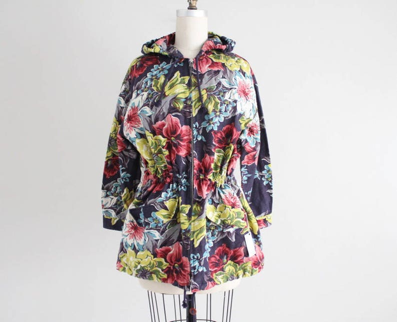 May include: A hooded jacket with a floral print in shades of pink, green, yellow, and blue. The jacket has a drawstring waist and a zipper closure.