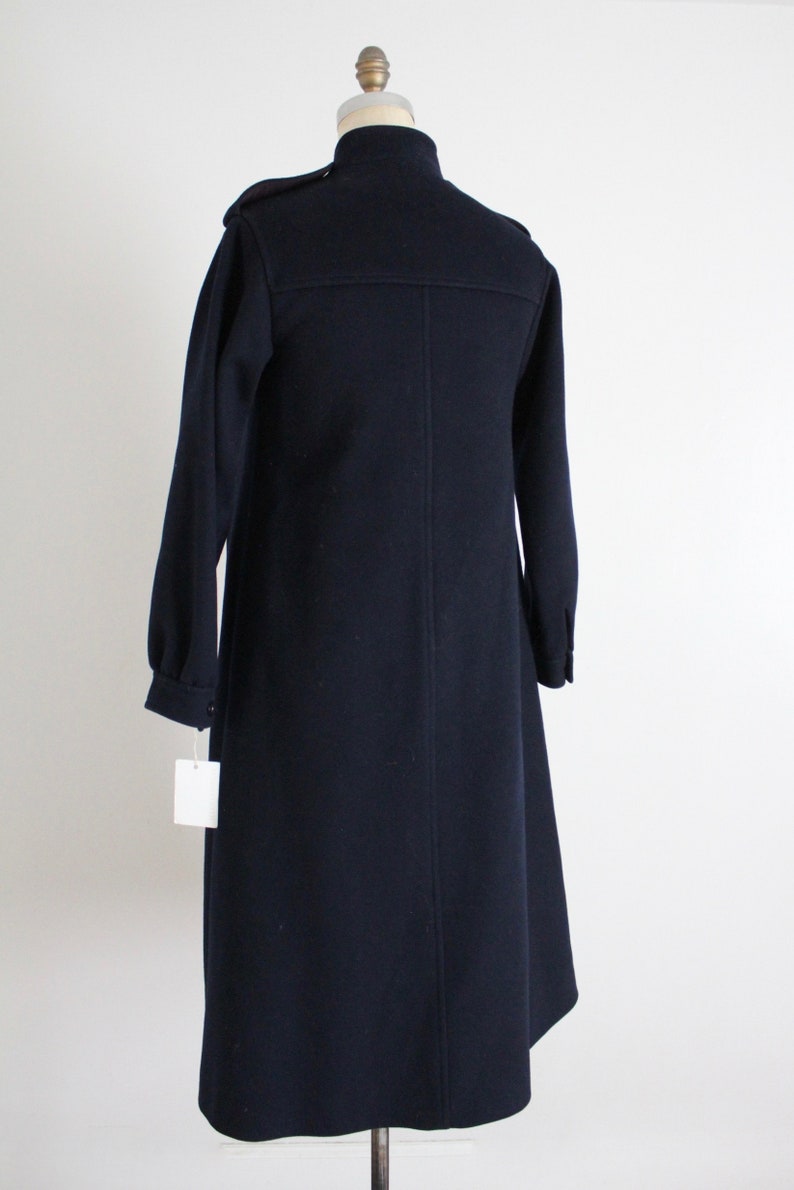 May include: A long, dark blue coat with a high collar and a single button closure. The coat has a slightly flared hem and is made of a thick, textured fabric.