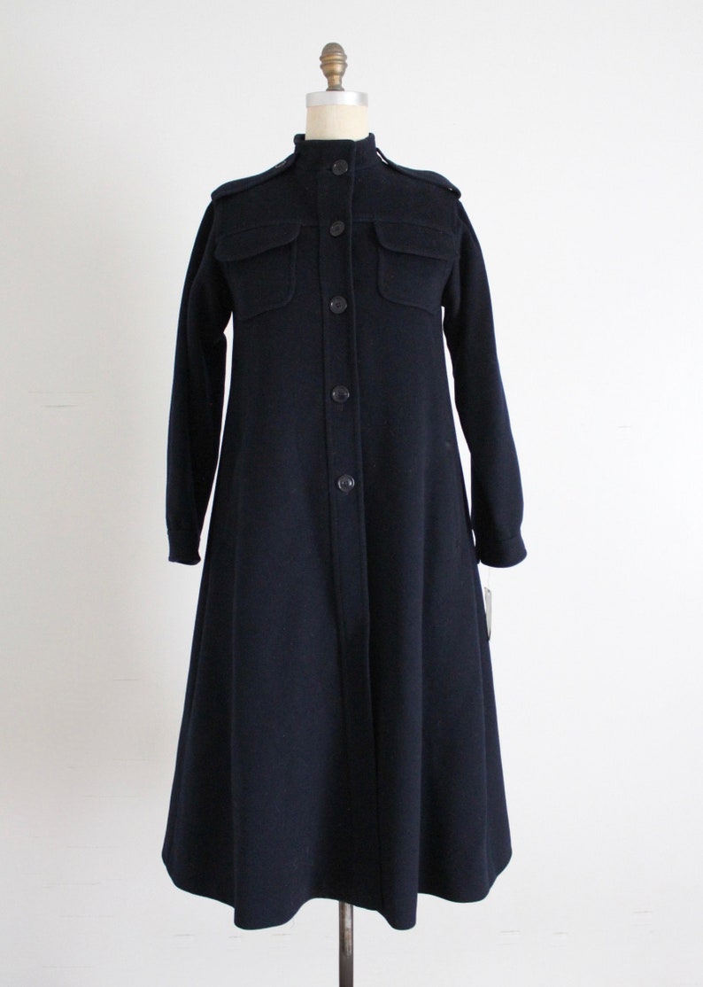 May include: A long, navy blue coat with a button-up front and large pockets. The coat has a stand-up collar and a slightly flared skirt.