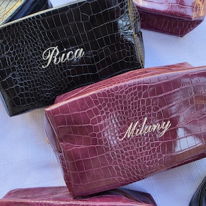 May include: A collection of rectangular cosmetic bags featuring a glossy crocodile-embossed texture. The bags are available in black and deep burgundy colors. Two of the bags are personalized with white embroidered names, specifically Rica and Milany.