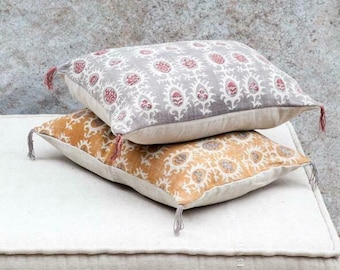 May include: Two decorative rectangular pillows stacked on top of each other. The top pillow is gray with a white and red floral pattern. The bottom pillow is mustard yellow with a white and brown floral pattern. Each pillow has tassels.
