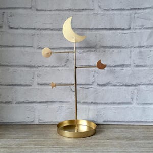 May include: A gold-colored jewelry stand with celestial shapes. The stand features a crescent moon, a star, and a circular shape. The base is a round tray. The stand is made of metal and is displayed against a white brick wall.