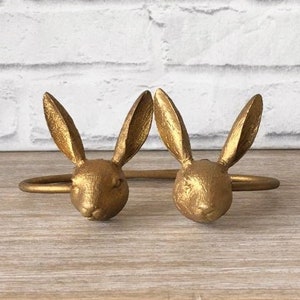 May include: Two gold-colored bunny head napkin rings. The bunny heads are detailed with textured fur and long ears. The rings are made of metal and are designed to hold napkins. The background is a white brick wall.