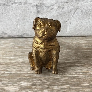 May include: A small, gold-coloured pug dog figurine. The pug is sitting upright with its front paws together. The figurine has detailed features, including wrinkles and a slightly upturned nose. The pug is made of a metallic material and has a shiny finish.