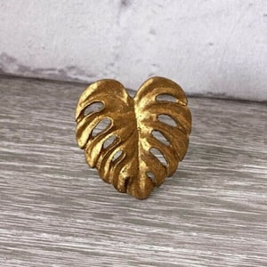 Puede incluir: Una hoja de metal de color dorado en forma de corazón con un diseño detallado. La hoja es un motivo de planta tropical.
