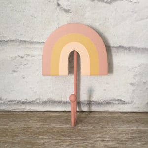 May include: A pink rainbow-shaped wall hook with yellow and cream stripes. The hook is made of metal and has a rounded end.