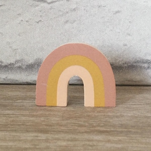 Può includere: Una decorazione arcobaleno in legno con tre archi in tonalità rosa, giallo e crema. L'arcobaleno è in piedi su una superficie di legno.