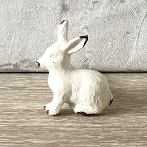 May include: A white ceramic rabbit ornament with black detailing on the ears and feet. The bunny is depicted in a seated pose, with its ears erect. The figurine has a slightly aged appearance, suggesting a vintage aesthetic.