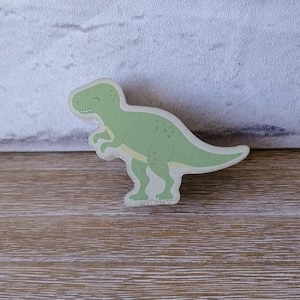 以下が含まれることがあります： 笑顔の顔をした緑色の木製恐竜のおもちゃ。恐竜は薄い茶色の木製の表面に立っています。