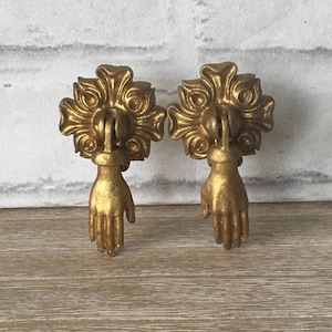 May include: A pair of antique gold-coloured drawer pulls. Each pull features a hand grasping a decorative floral element. The pulls are made of metal and have a distressed finish, giving them a vintage appearance. The background is a white brick wall.