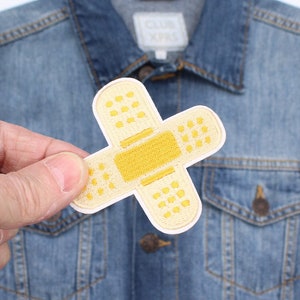 May include: An embroidered bandage patch in yellow and white, held against a denim jacket. The patch features a yellow central pad and white edges with small yellow dots, a fun accessory.