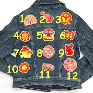 May include: A blue denim jacket decorated with embroidered patches. Patches include a rainbow, guitar, peace sign, flower, VW bus, butterfly, mushroom, and a smiling flower. The jacket has a button closure.