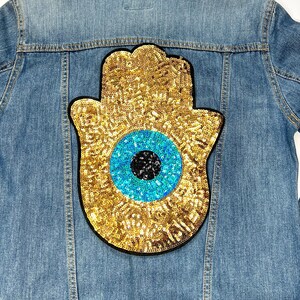 以下が含まれることがあります： 背中に大きな金色のスパンコールで飾られたハムサハンドパッチが付いた、ライトブルーのデニムジャケット。 パッチは黒い瞳を持つ青い目があります。 ジャケットはライトブルーウォッシュです。