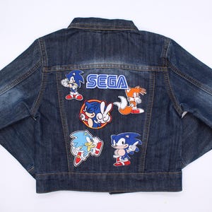 May include: A dark blue denim jacket with various colourful Sonic the Hedgehog character patches and the SEGA logo on the back. Patches include Sonic, Tails, and other characters from the video game series.