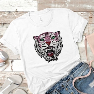 May include: White t-shirt with a sequined tiger head design. The tiger has a pink and silver face, green eyes, and a red nose. The shirt is styled with denim shorts, sandals, and a pink handbag.