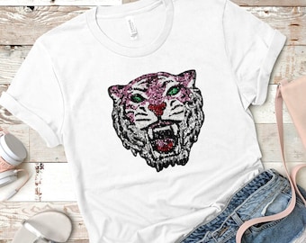 以下が含まれることがあります： スパンコールで装飾された虎の頭のデザインが特徴の白いTシャツ。虎はピンクとシルバーの顔、緑の目、赤い鼻をしています。このシャツは、デニムショーツ、サンダル、ピンクのハンドバッグと組み合わせています。